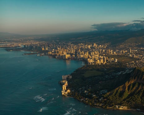 Sunset view of edge of volcanic crater, with high rise buildings along a beach, and blue ocean.