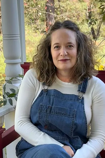 A woman wearing overalls and a white shirt smiles at the camera, seated outside on a bench.