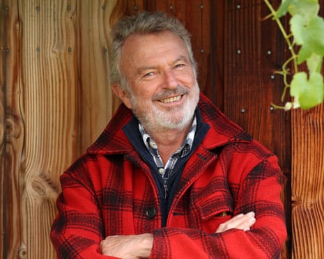 Actor Sam Neill smiling with arms crossed in a red plaid jacket in front of a wooden fence