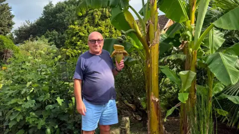Supplied A man wearing glasses, a dark blue T-shirt and light blue shorts stands next to a banana plant. There are a number of trees and plants behind him.