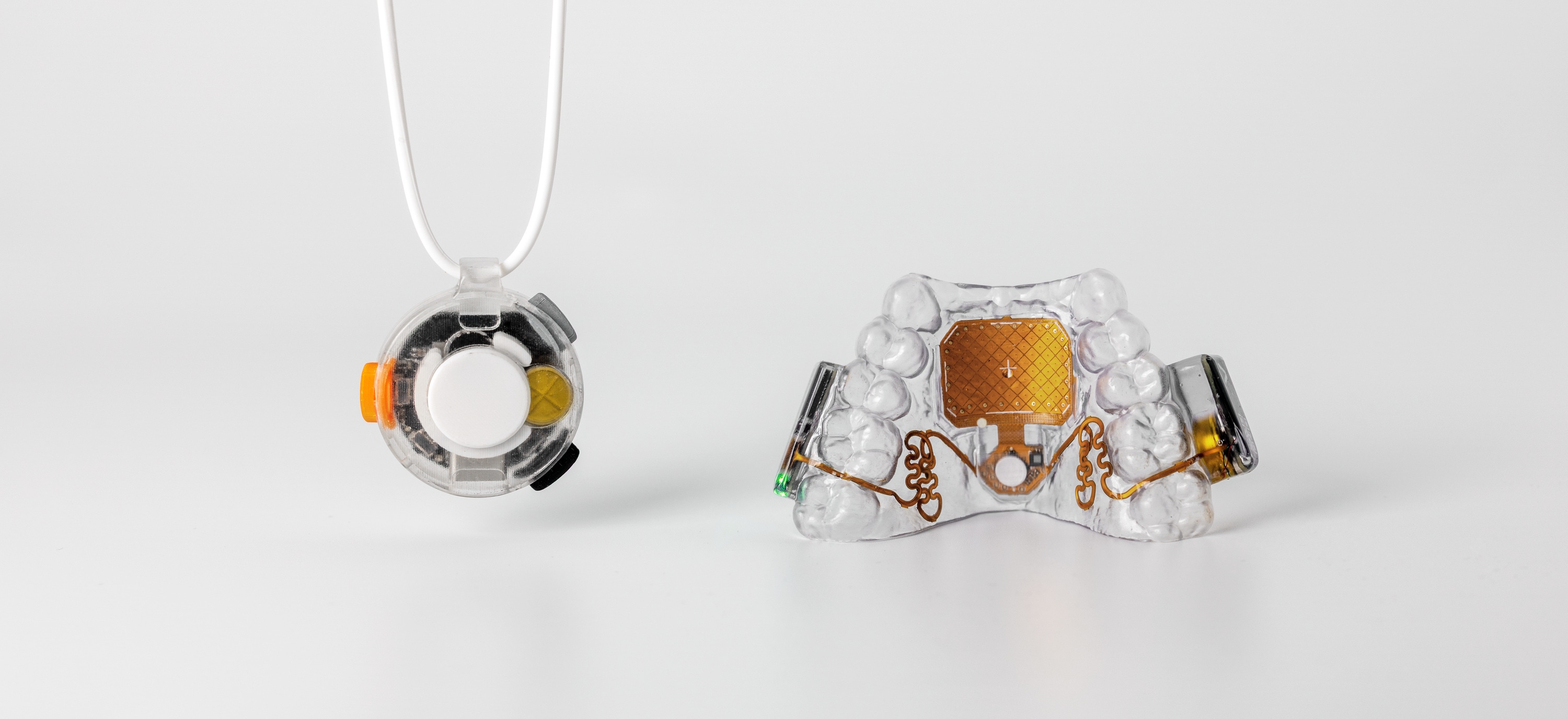 VOX and the MouthPad side by side on a pale studio background. VOX hangs at the left on its cord: a small round device in a clear housing, its orange side control and pale capture button visible through the case. The MouthPad sits at the right: a clear custom-fit retainer with a gold touchpad at its centre and gold circuitry curving out to the side electronics.