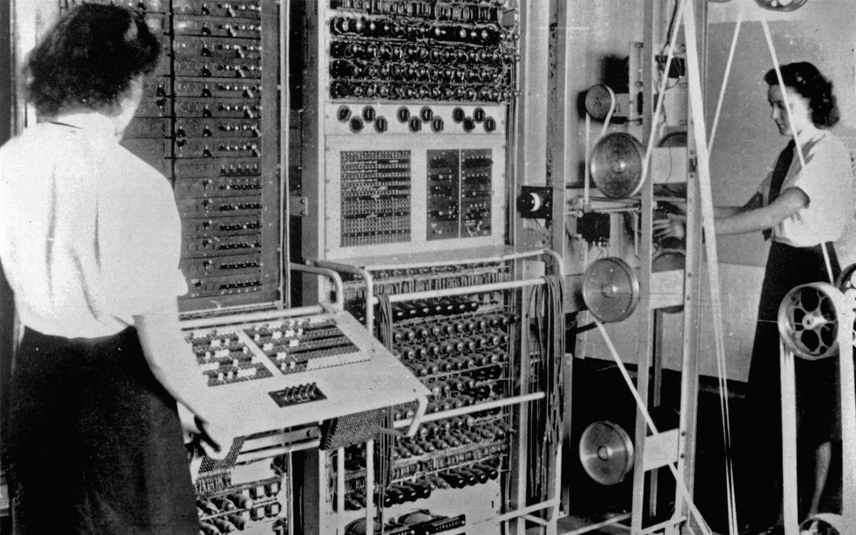 Black and white image of two women operating a wall-sized 1940s computer.