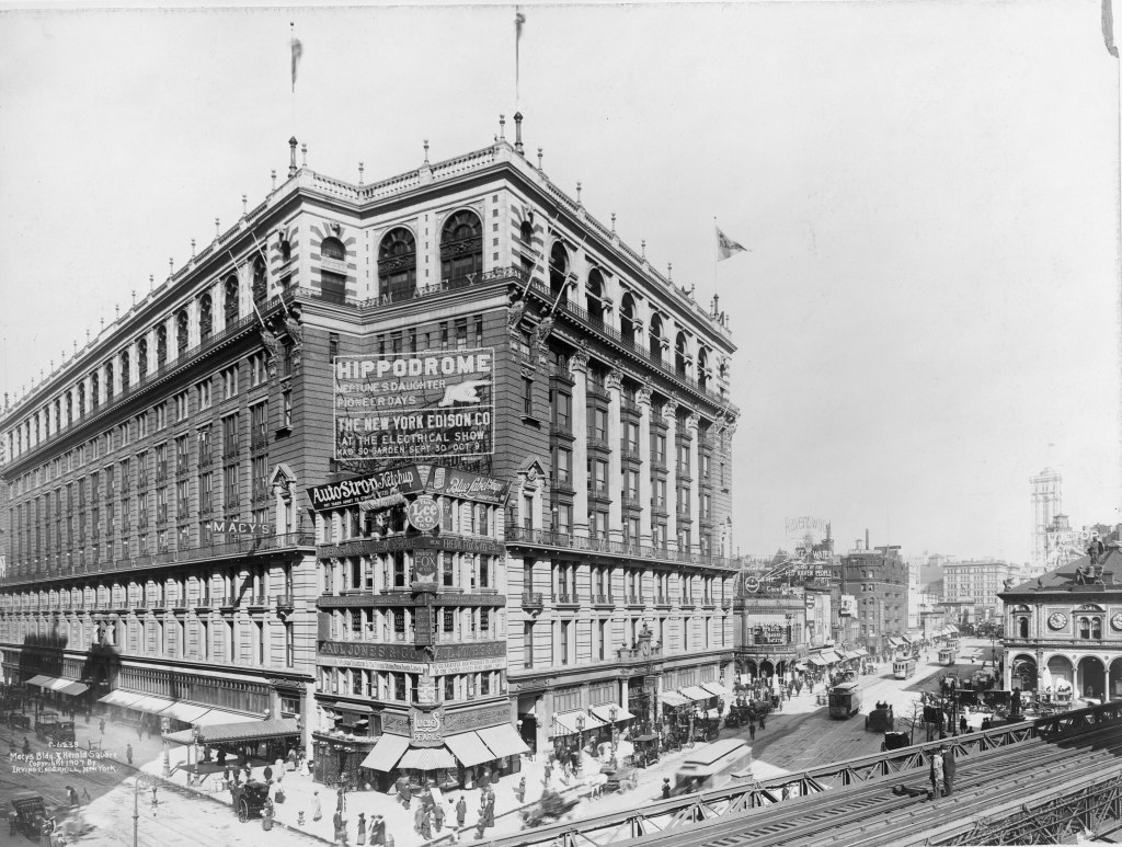 Historic black and white photograph of the Hippodrome Theater building in New York, featuring banners for performances and advertisements, with bustling street activity and vintage vehicles in the foreground.