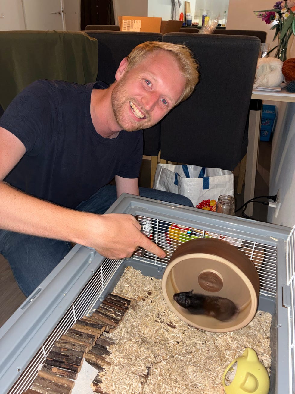 A man points excitedly at a hamster running in its wheel inside a small pet cage.