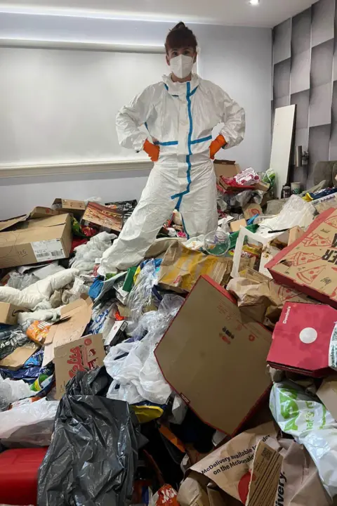 Supplied A woman in white overalls and orange gloves as well as a white mask is standing in a room full of cardboard and rubbish. She has her hands on her hips and her dark hair is tied up.