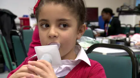 BBC Young child with brown hair, blowing air into plastic measuring device. She wears a red ribbon and red cardigan and a white shirt, She is in school, sitting on a green chair. 