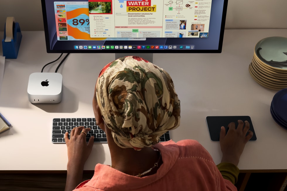 An overhead view of a person using a keyboard and mouse at a desk, with a Mac mini connected to a large display.