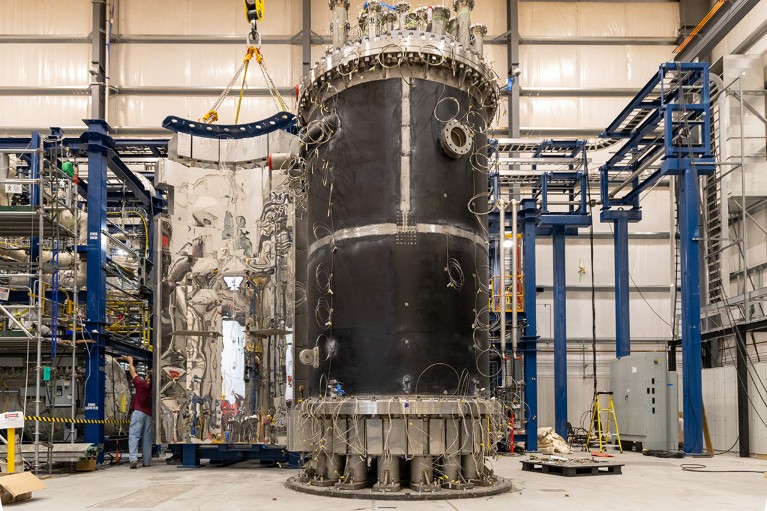 A large reactor vessel is in the centre of a room full of equipment. A person can be seen to the right hand side of the room.