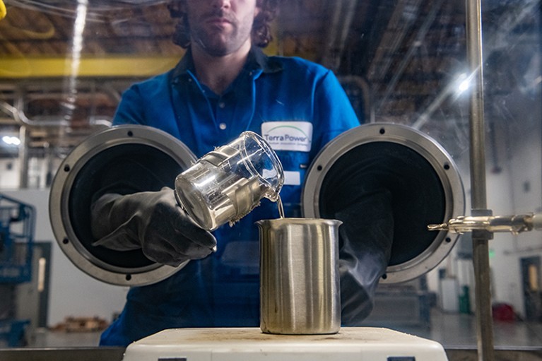 Person wearing blue overalls seen through glass. Person is wearing black protective gloves and pouring liquid from one container to another.