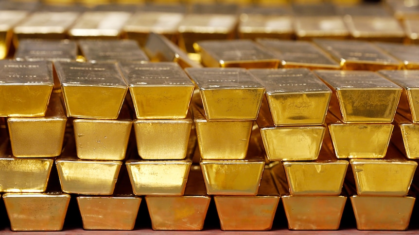 The ends of gold bullion bars, seen stacked four rows high and six wide, in front of other blurred piles of gold.
