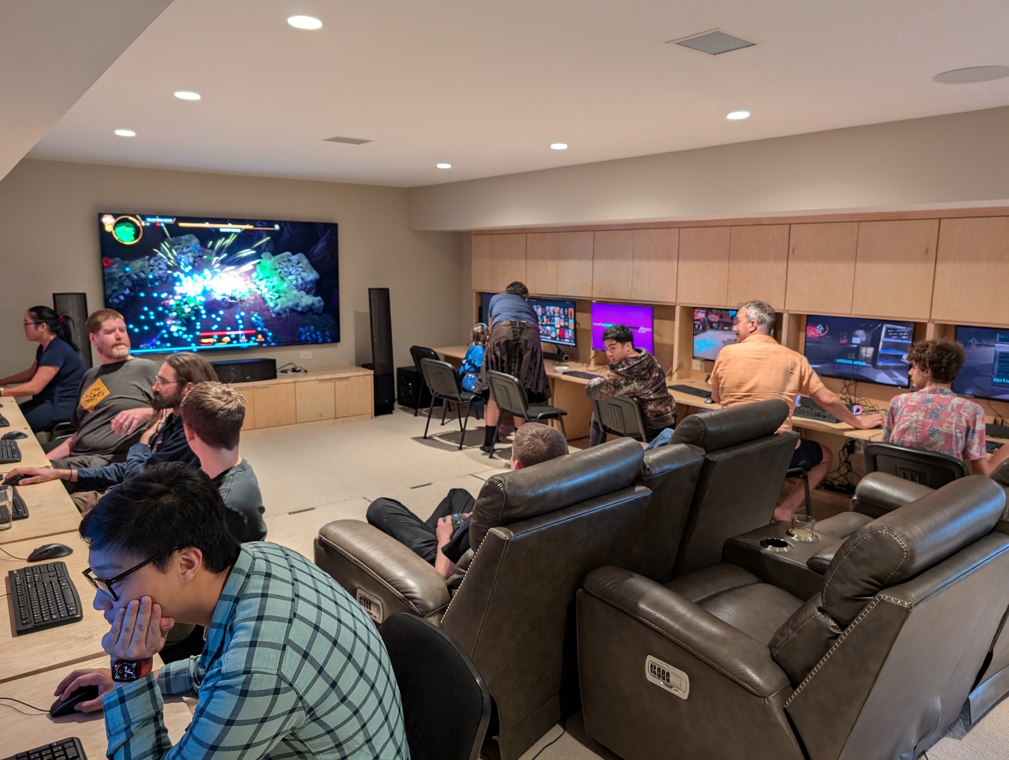 A photo depicts the game room, 18 feet wide and 24 feet long, full of people playing video games. The room is a finished basement room; it is carpeted with beige walls and no windows. On the far wall is a 98-inch TV. Along both side walls are rows of computer screens, six on each side. Each screen sits inside wood panel cabinetry which folds out to provide a desk in front of it. People are sitting at all the desks, playing video games. In the middle of the room, facing the TV are two recliner loveseats. Someone is sitting at one of them, holding a controller and playing a video game on the TV.