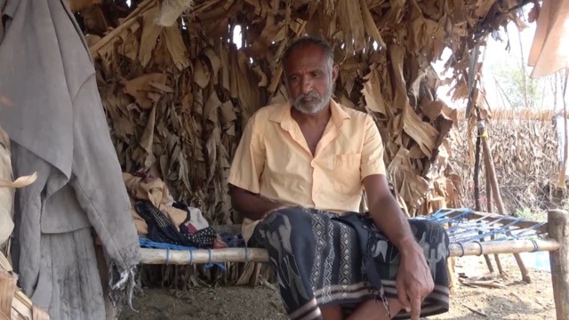 A wide shot of a man sitting on a makeshift bed under a makeshift shelter in an arid desert landscape. He has a grey beard and short darker hair and wears a yellow shirt, with a robe-like garment covering his legs. He has a neutral expression and is looking towards the ground away from the camera