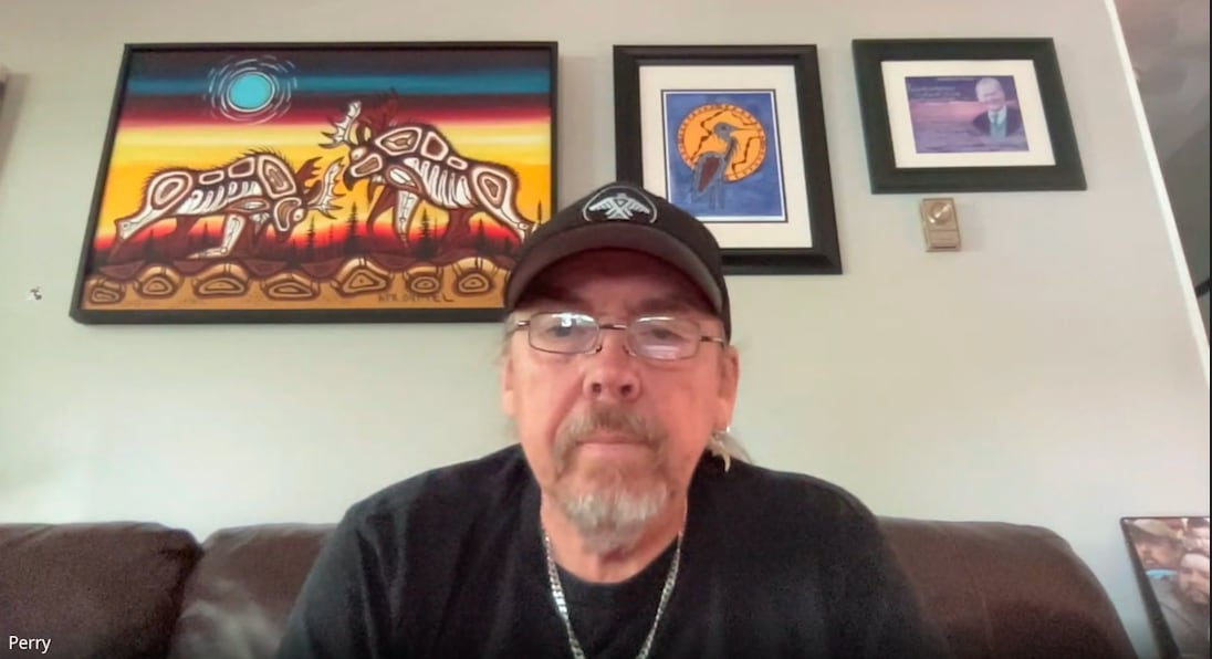 A man with a hat and glasses and a number of framed pictures behind him.
