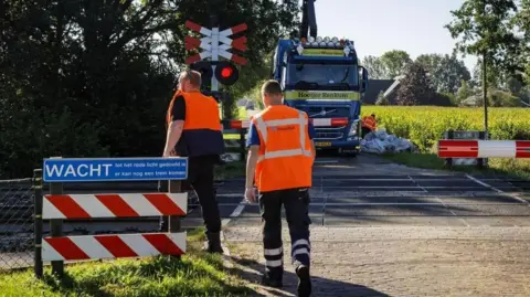 EPA Rail workers in hi vis jackets are pictured at a level crossing in Holten in the Netherlands on Tuesday. In the background a lorry can be seen parked on the other side of the crossing, while another worker is seen next to a large pile of building sacks.