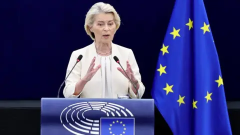 EPA Ursula von der Leyen, who has a grey/blonde short haircut, wears a cream blazer and blouse as she speaks at a podium, with the EU flag in the background